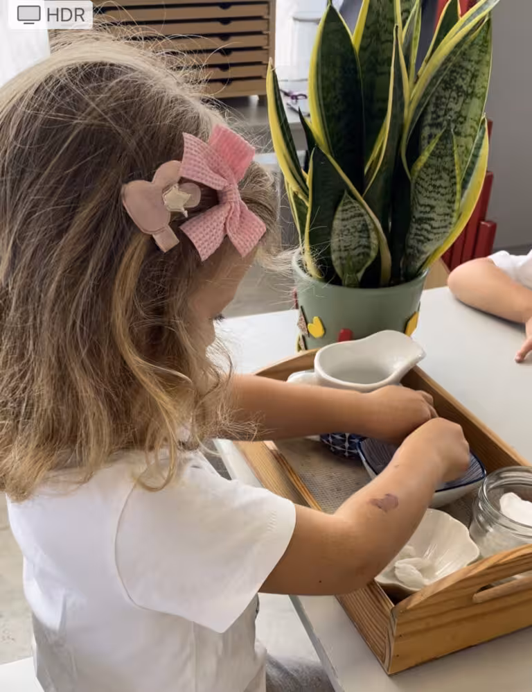 Niña del IMS Sotogrande con lazo rosa trabajando con bandeja de vida práctica Montessori junto a planta